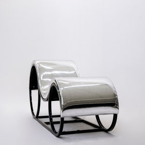 May include: A modern chaise lounge with a reflective silver seat and black metal frame. The curved design features two cushioned sections connected by a rectangular base and rounded supports, creating a sleek, minimalist aesthetic.