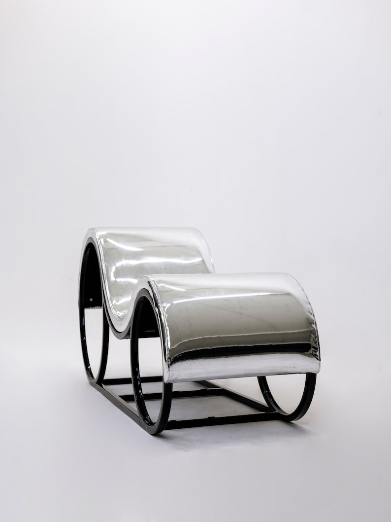 May include: A modern chaise lounge with a reflective silver seat and black metal frame. The curved design features two cushioned sections connected by a rectangular base and rounded supports, creating a sleek, minimalist aesthetic.