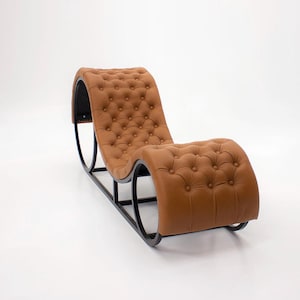 May include: A modern chaise lounge chair with a tufted, caramel-colored leather seat and backrest. The chair has a curved design and a black metal frame. The overall aesthetic is sleek and contemporary, suitable for a living room or lounge area.