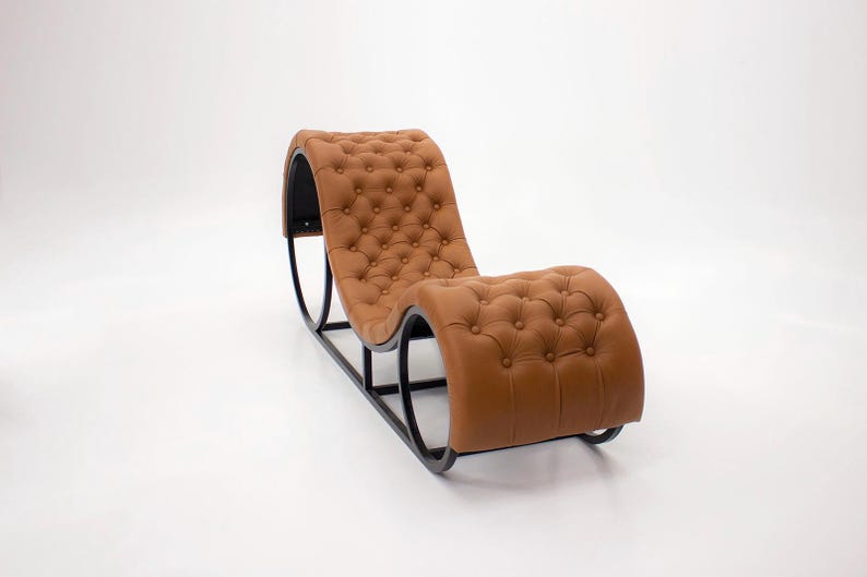 May include: A modern chaise lounge chair with a tufted, caramel-colored leather seat and backrest. The chair has a curved design and a black metal frame. The overall aesthetic is sleek and contemporary, suitable for a living room or lounge area.
