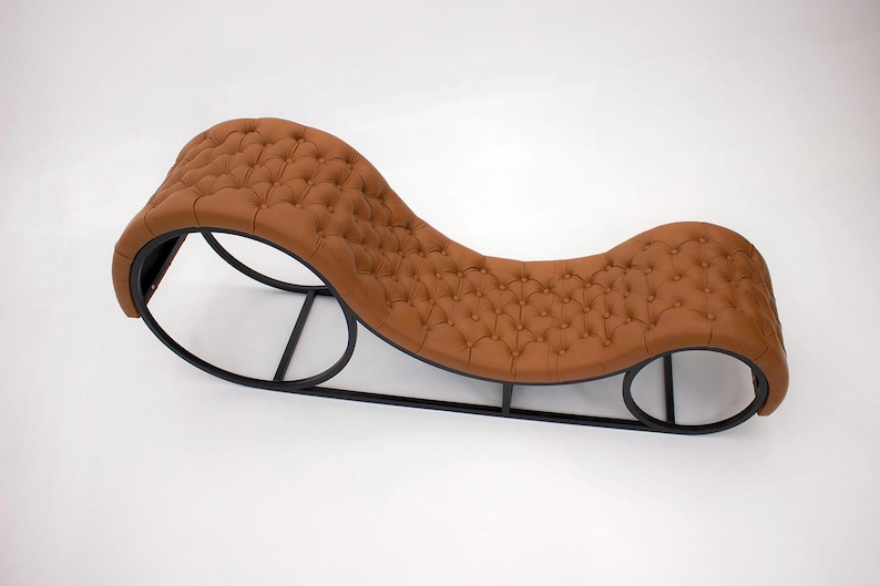 May include: A brown chaise lounge with a tufted seat and backrest. The curved design features a black metal frame. The lounge is designed for relaxation and adds a touch of elegance to any living space.