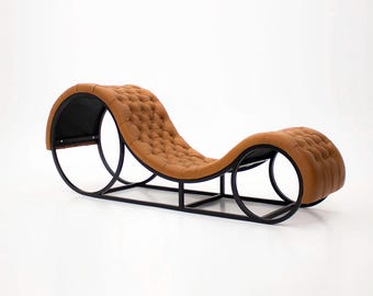 May include: A modern chaise lounge with a tufted, caramel-colored leather seat. The seat is supported by a black metal frame with curved, circular elements at each end. The design is sleek and minimalist, suitable for contemporary interiors.