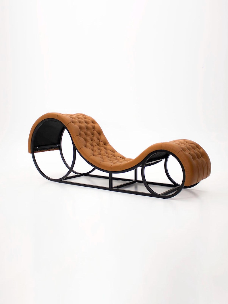 May include: A modern chaise lounge with a tufted, caramel-colored leather seat. The seat is supported by a black metal frame with curved, circular elements at each end. The design is sleek and minimalist, suitable for contemporary interiors.