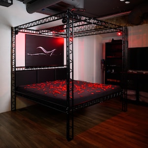 May include: A black metal four-poster bed frame with a truss-style design. The bed features a dark mattress covered in scattered red rose petals. A large rectangular black and white abstract art piece is mounted on the wall behind the headboard.