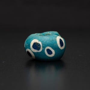 May include: A turquoise glass bead with white and dark blue circular patterns. The bead has a cylindrical shape with a hole through the centre. The bead is likely an antique or vintage item.