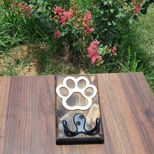 May include: A wooden wall hook with a paw print cutout and two black metal hooks. The wood has a dark stain with a lighter paw print shape. The hook is designed for pet lovers.