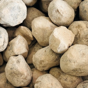 May include: A close-up of a pile of smooth, light brown stones. The stones are various sizes and shapes, and they have a slightly rough texture.