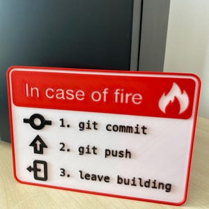 Może przedstawiać: Czerwono-biały znak z napisem "W razie pożaru" i ikoną płomienia. Znak zawiera trzy kroki: 1. git commit, 2. git push, 3. opuść budynek.