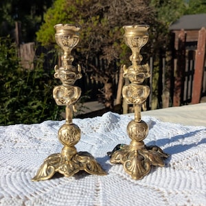 May include: A pair of matching brass candlesticks featuring a central figure with horns and pointed ears. Each candlestick has a wide, ornate base with decorative scrollwork, a spherical midsection, and a cup at the top for holding a candle.
