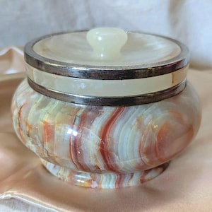 May include: A decorative jar with a lid, crafted from a blend of cream, orange, and brown onyx. The jar features a silver-toned band around the lid and base, and a small, rounded knob on top. The jar is set on a satin surface.