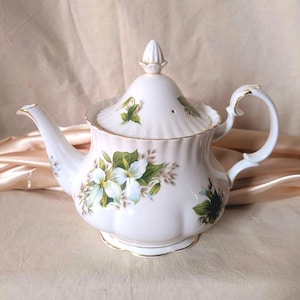 May include: A white porcelain teapot featuring a scalloped body and a matching lid with a pointed finial. The teapot is decorated with painted white trillium flowers, green leaves, and gold trim along the edges, handle, and spout base.