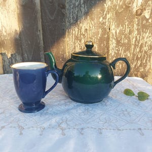 May include: A dark green teapot with a gold trim and a matching blue teacup. The teapot and teacup are sitting on a white tablecloth with a delicate pattern.