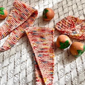 Puede incluir: Tres bufandas de crochet rosa y blanco con un patrón de hilo multicolor. Las bufandas están dispuestas sobre una superficie de crochet blanco con fresas rosas pintadas.