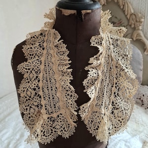 May include: Antique cream-colored lace collar with a delicate, openwork design. The collar is displayed on a vintage brown dress form, showcasing its intricate floral and geometric patterns. The lace has a scalloped edge.