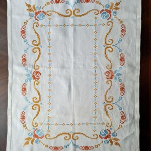 May include: White linen table runner with a detailed cross-stitch design. The embroidery features a repeating pattern of orange, blue, and gold floral and scroll motifs, creating a decorative border.