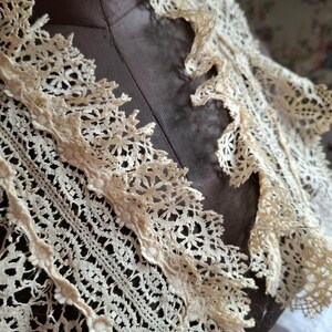 May include: Close-up of an antique cream-colored lace collar. The collar features intricate floral and geometric patterns, with a scalloped edge. The lace is delicate and appears to be handmade, showcasing a vintage aesthetic. The collar is displayed on a mannequin.