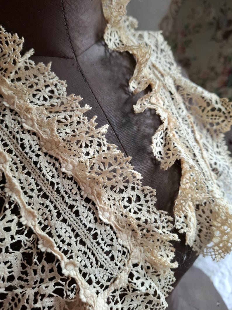 May include: Close-up of an antique cream-colored lace collar. The collar features intricate floral and geometric patterns, with a scalloped edge. The lace is delicate and appears to be handmade, showcasing a vintage aesthetic. The collar is displayed on a mannequin.