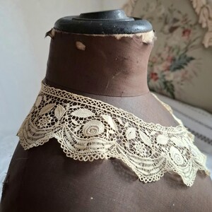 May include: An antique cream-colored lace collar with floral and leaf patterns. The delicate lace is displayed on a brown mannequin head. The collar's intricate design is visible against the dark background, showcasing its vintage charm.