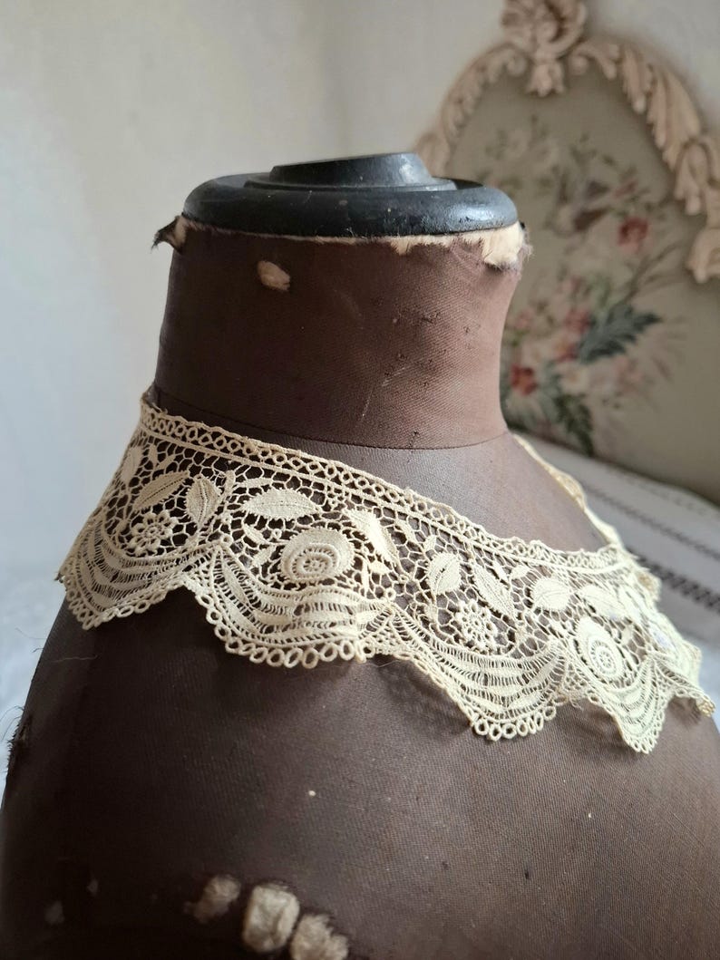 May include: An antique cream-colored lace collar with floral and leaf patterns. The delicate lace is displayed on a brown mannequin head. The collar's intricate design is visible against the dark background, showcasing its vintage charm.
