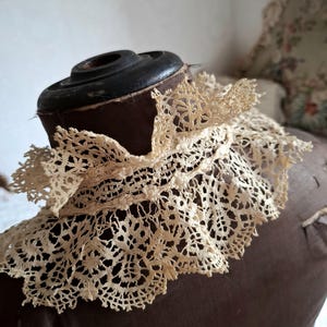 May include: An antique cream-colored lace collar, featuring an intricate floral pattern. The delicate lace is draped over a brown dress form, showcasing its detailed openwork design. The collar's edges are scalloped, adding to its vintage charm.