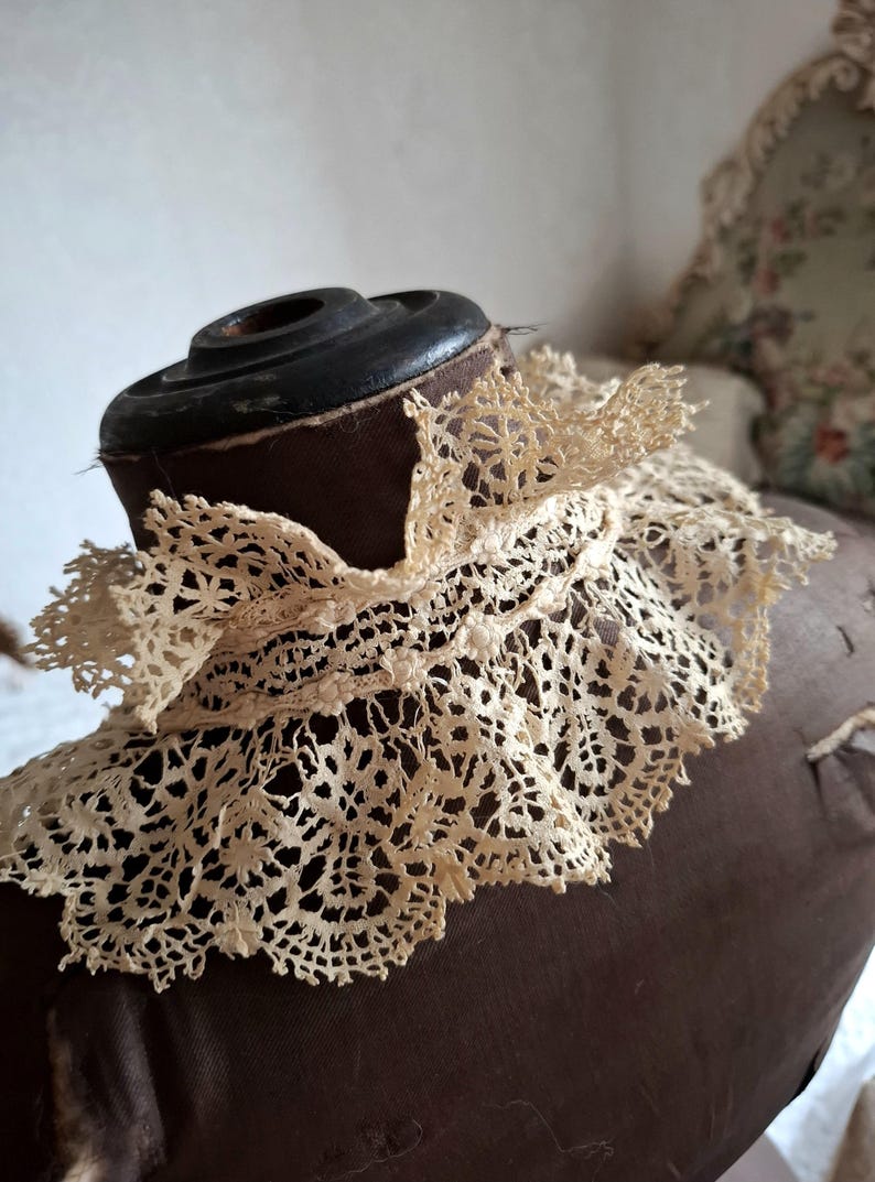 May include: An antique cream-colored lace collar, featuring an intricate floral pattern. The delicate lace is draped over a brown dress form, showcasing its detailed openwork design. The collar's edges are scalloped, adding to its vintage charm.