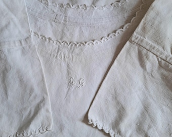 May include: A white linen garment with scalloped edges and embroidered initials "L.C." on the chest. The sleeves are wide and the neckline is detailed with a delicate trim. The fabric appears to be soft and slightly textured.
