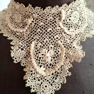 May include: Cream-colored, antique lace collar with intricate openwork and floral patterns. The collar is displayed on a dark brown mannequin, showcasing its delicate design and vintage aesthetic. The lace features a scalloped edge and detailed circular motifs.