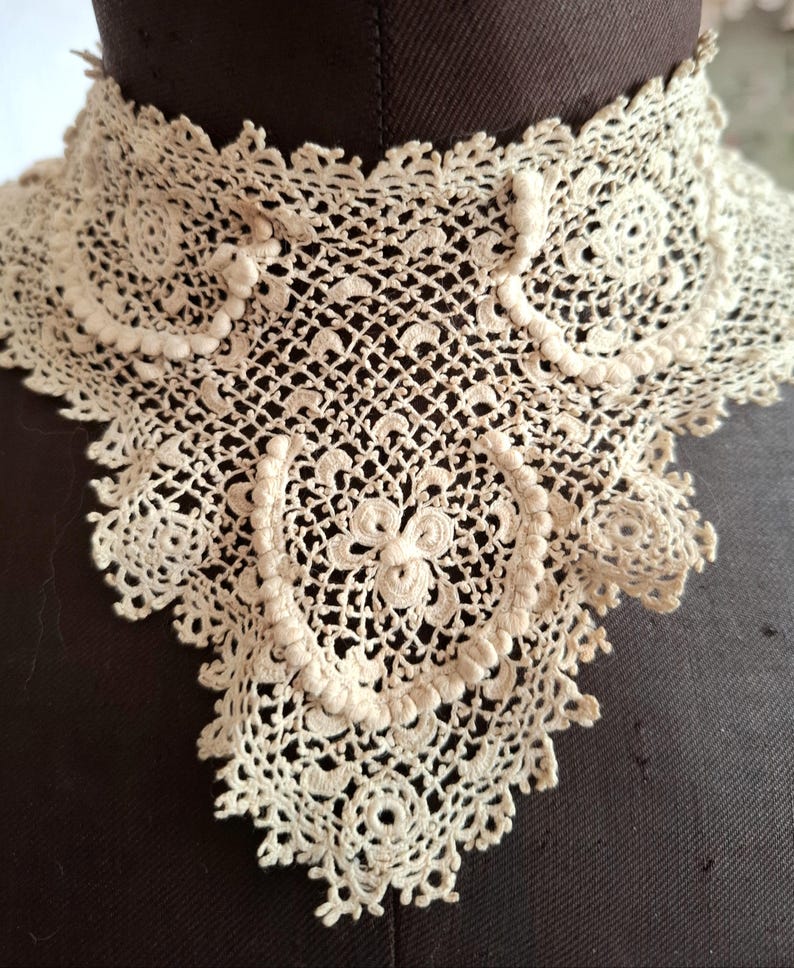 May include: Cream-colored, antique lace collar with intricate openwork and floral patterns. The collar is displayed on a dark brown mannequin, showcasing its delicate design and vintage aesthetic. The lace features a scalloped edge and detailed circular motifs.