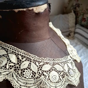 May include: Cream-colored lace collar with a floral and leaf design. The collar is displayed on a brown dress form. The lace features intricate patterns and a scalloped edge, showcasing a vintage aesthetic.