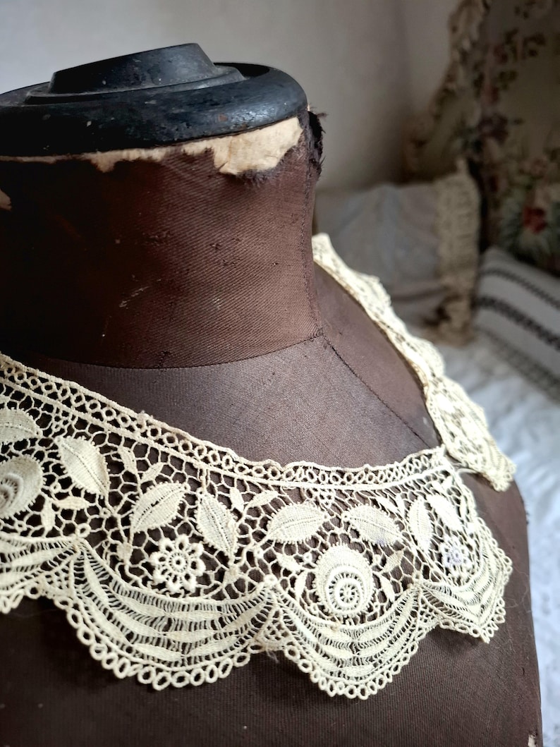 May include: Cream-colored lace collar with a floral and leaf design. The collar is displayed on a brown dress form. The lace features intricate patterns and a scalloped edge, showcasing a vintage aesthetic.