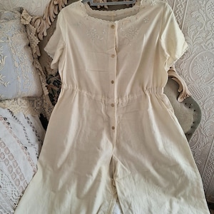May include: A cream-colored short-sleeve romper featuring a button-down front, a cinched elastic waist, and a scalloped neckline. The garment is decorated with delicate white floral embroidery on the chest and along the scalloped leg hems.