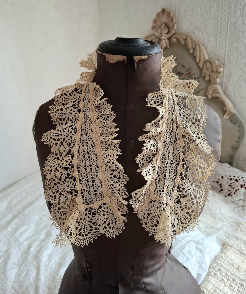 May include: Antique cream-colored lace collar with a delicate, openwork design. The collar is displayed on a vintage brown dress form, showcasing its intricate floral and geometric patterns. The lace has a scalloped edge.