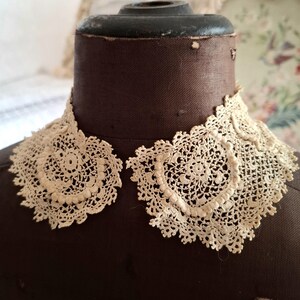 May include: Cream-colored, antique lace collar with a delicate, openwork design. The collar is displayed on a brown dress form, showcasing its intricate floral and geometric patterns. The lace appears to be handmade, with a scalloped edge.