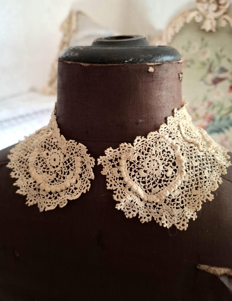 May include: Cream-colored, antique lace collar with a delicate, openwork design. The collar is displayed on a brown dress form, showcasing its intricate floral and geometric patterns. The lace appears to be handmade, with a scalloped edge.