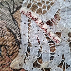 May include: Close-up of antique white lace with a delicate, openwork design. A small, rectangular tag embroidered with the name "Freda Cook" and the year "1899" in red thread is attached to the lace. The lace is set against a patterned background.