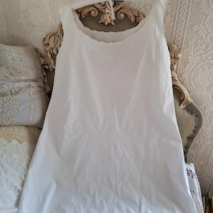 May include: A sleeveless white slip dress featuring a scoop neckline with delicate white floral embroidery around the collar. The garment has a simple, straight-cut silhouette and a plain hemline, presented on a padded hanger for display.