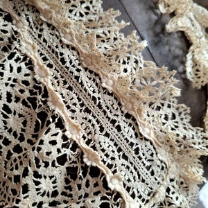 May include: Close-up of antique cream-colored lace with intricate floral and geometric patterns. The lace features delicate scalloped edges and a detailed openwork design, showcasing the craftsmanship of the textile.