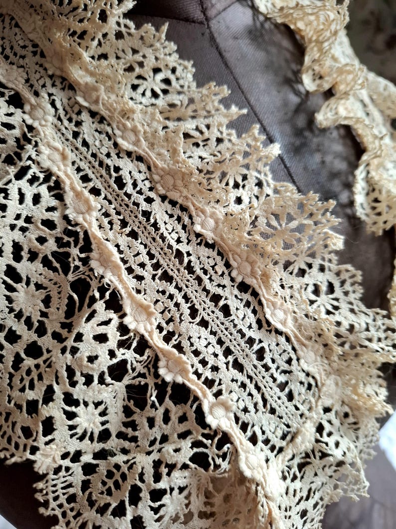 May include: Close-up of antique cream-colored lace with intricate floral and geometric patterns. The lace features delicate scalloped edges and a detailed openwork design, showcasing the craftsmanship of the textile.