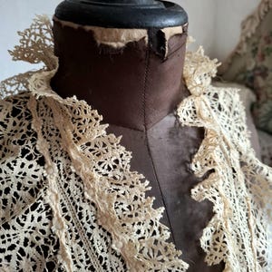 May include: A vintage cream-colored lace collar is displayed on a brown dress form. The lace features a delicate floral pattern and a scalloped edge. The dress form has a black top and is set against a neutral background.