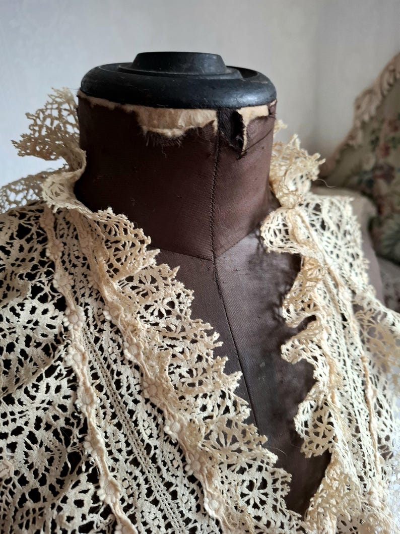 May include: A vintage cream-colored lace collar is displayed on a brown dress form. The lace features a delicate floral pattern and a scalloped edge. The dress form has a black top and is set against a neutral background.