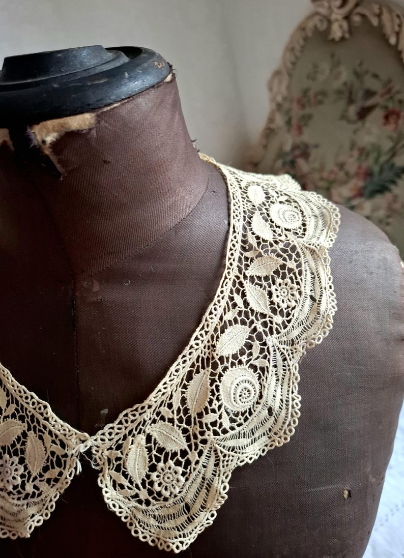 May include: Cream-colored lace collar with a floral and leaf pattern, displayed on a brown dress form. The collar features intricate openwork and a scalloped edge, showcasing a vintage aesthetic.