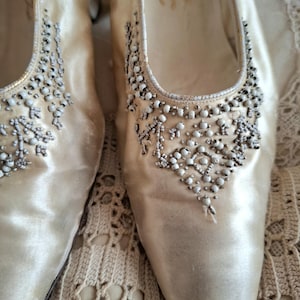 Może przedstawiać: Wytworne, kremowe buty vintage z satyny, zdobione koralikami. Buty mają szpiczasty nosek i niski obcas. Ozdoby z koralik&oacute;w skupione są wok&oacute;ł palc&oacute;w i na g&oacute;rnej części buta. Idealne na specjalną okazję.