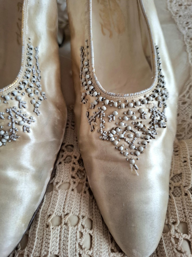 Może przedstawiać: Wytworne, kremowe buty vintage z satyny, zdobione koralikami. Buty mają szpiczasty nosek i niski obcas. Ozdoby z koralik&oacute;w skupione są wok&oacute;ł palc&oacute;w i na g&oacute;rnej części buta. Idealne na specjalną okazję.
