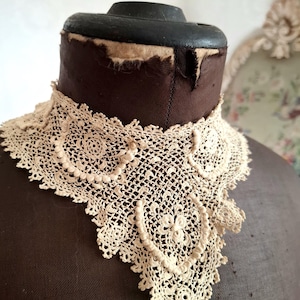 May include: Cream-colored, antique lace collar with a detailed, openwork design. The collar is displayed on a brown dress form, showcasing its intricate floral and geometric patterns. The lace is delicate and appears to be handmade.
