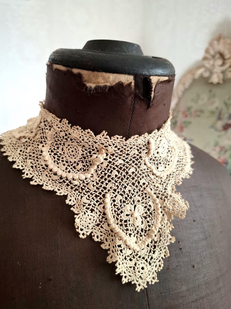 May include: Cream-colored, antique lace collar with a detailed, openwork design. The collar is displayed on a brown dress form, showcasing its intricate floral and geometric patterns. The lace is delicate and appears to be handmade.