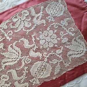 Könnte beinhalten: Nahaufnahme von antiker weißer Spitze mit floralen und scrollartigen Designs, die auf einem tiefrosafarbenen Stoff liegt. Die Spitze hat ein zartes, offenes Gewebe, und der Stoff hat einen subtilen Glanz. Das Bild deutet auf Vintage-Textilien hin.