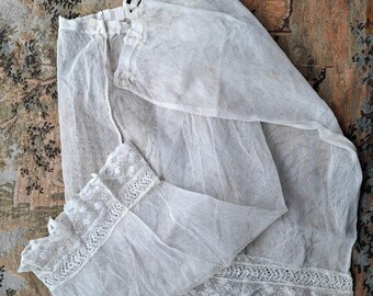 Pode incluir: Uma peça de roupa interior branca vintage, possivelmente uma anágua ou camisola, feita de tecido transparente. A peça apresenta delicadas bordas de renda nas bordas e um decote franzido. O tecido é ligeiramente translúcido e a peça está dobrada.