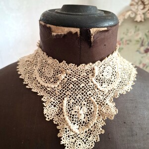May include: An antique cream-colored lace collar, displayed on a brown dress form. The collar features intricate floral and geometric patterns, with a scalloped edge. The delicate lace is a vintage textile.