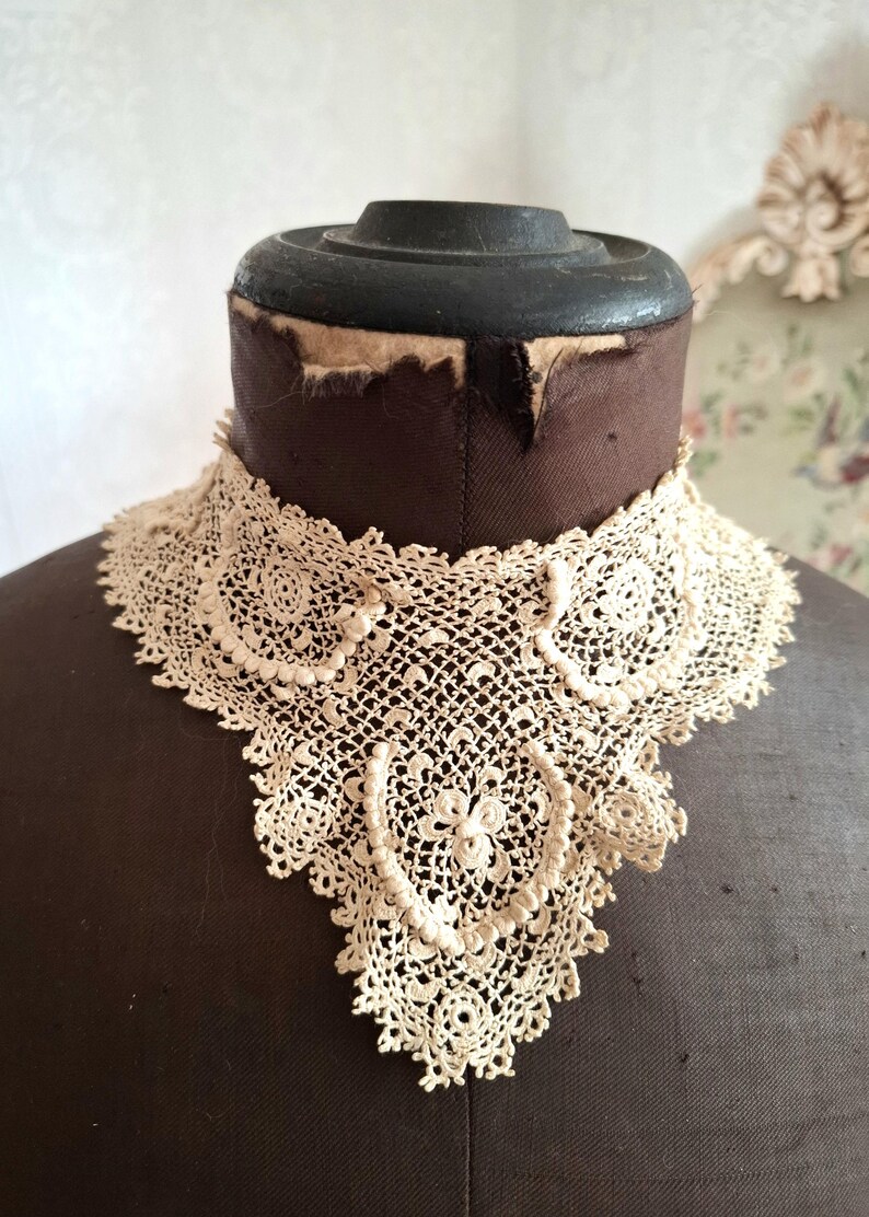 May include: An antique cream-colored lace collar, displayed on a brown dress form. The collar features intricate floral and geometric patterns, with a scalloped edge. The delicate lace is a vintage textile.