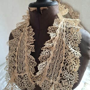 May include: An antique cream-colored lace collar with intricate floral and geometric patterns. The collar is draped over a brown mannequin, showcasing its delicate, openwork design and ruffled edges. The vintage textile adds a touch of elegance.